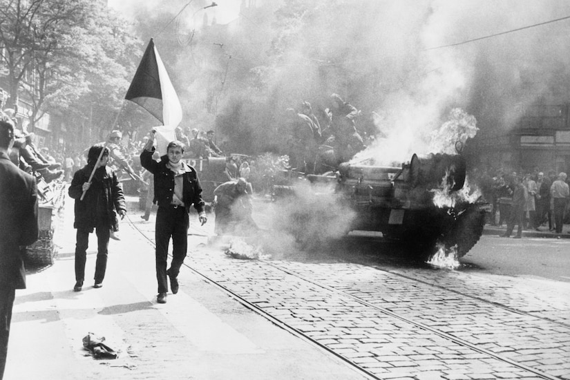 A war scene shows a tank burning in the street and citizens with a flag running along the sidewalk.
