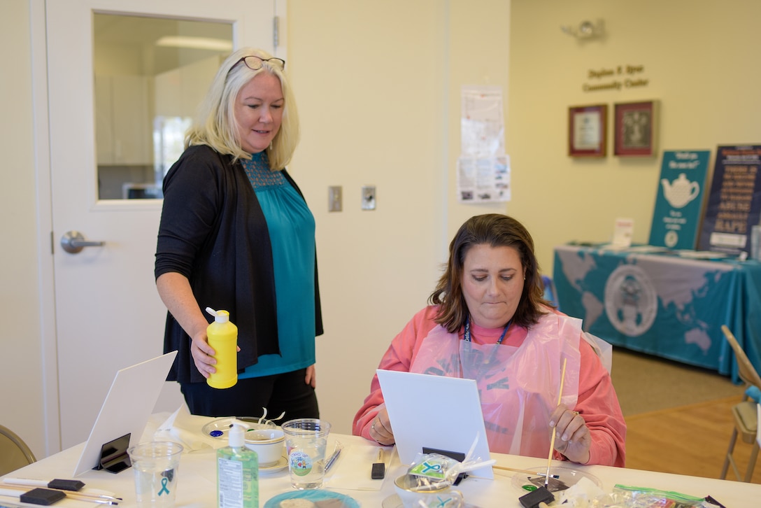 A woman offers yellow paint to a female participant dressed in pink sitting at a table painting her own painting.