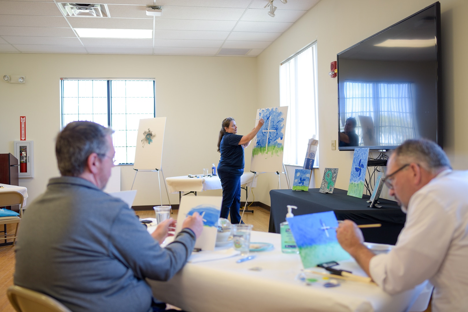 A woman at an easel paints a painting. Several people sitting at tables paint their own paintings with her.