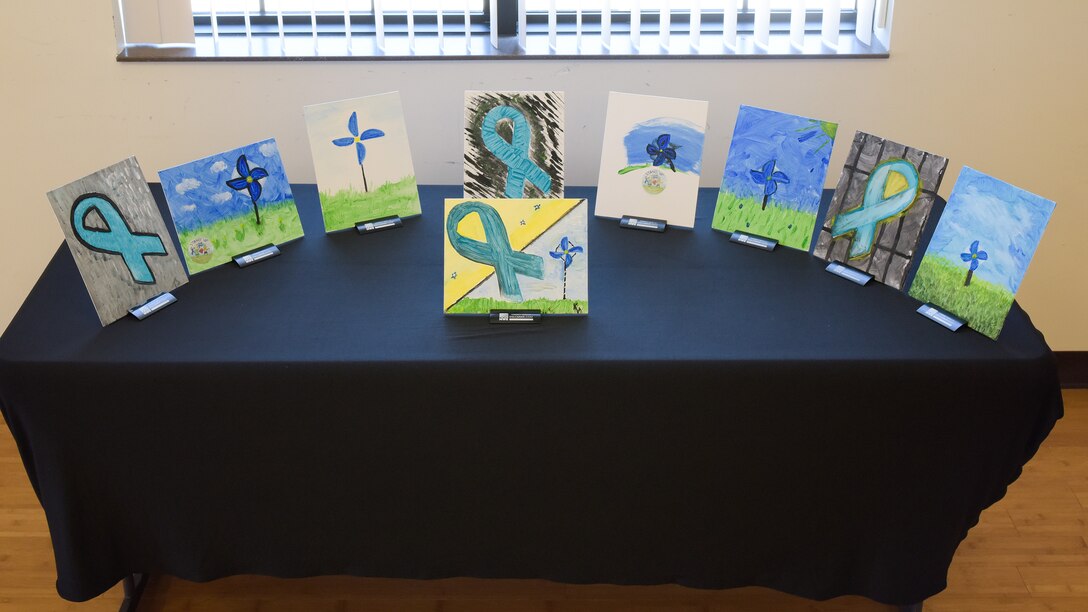Nine painted canvases on a table with a black tablecloth. Paintings have a mix of teal ribbons and blue pinwheels on them.
