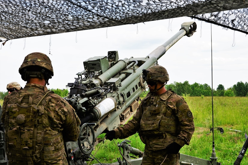 Soldiers fire a large gun as they stand under a camouflage net.