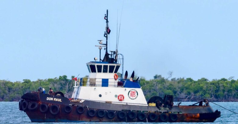 Black and white tugboat on a blue waterway with green trees in the background.