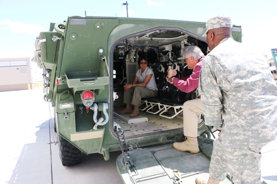 A woman sits in the back of a combat vehicle. Two men stand near the door of the vehicle.