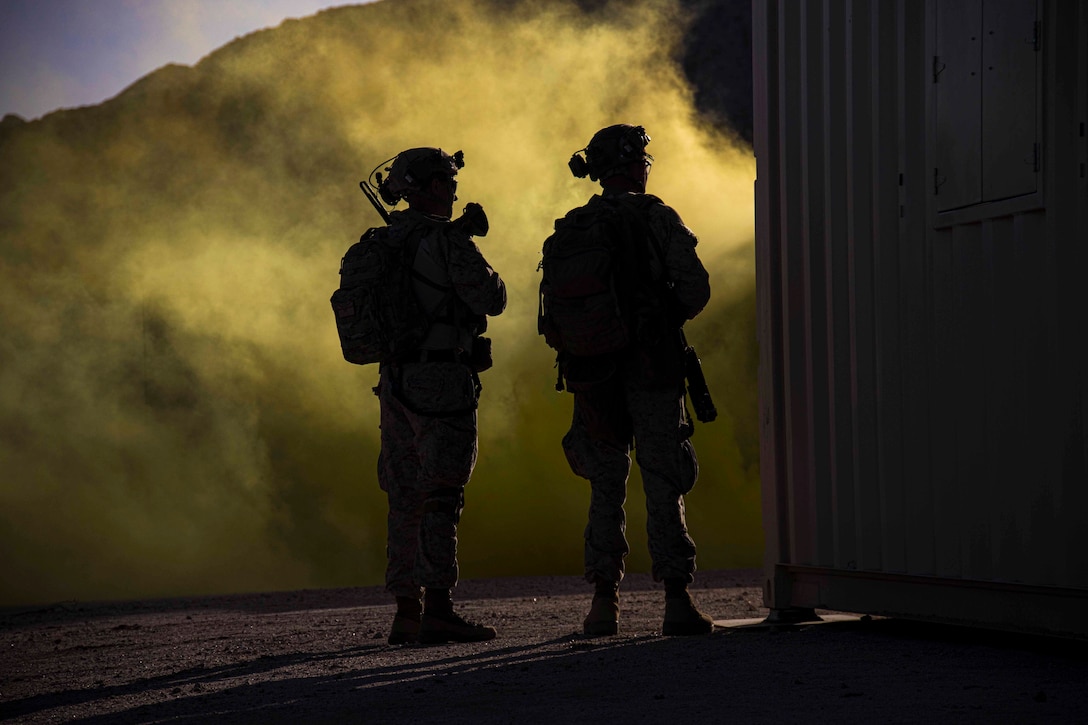 Two Marines shown in silhouette stand in front of yellow smoke.