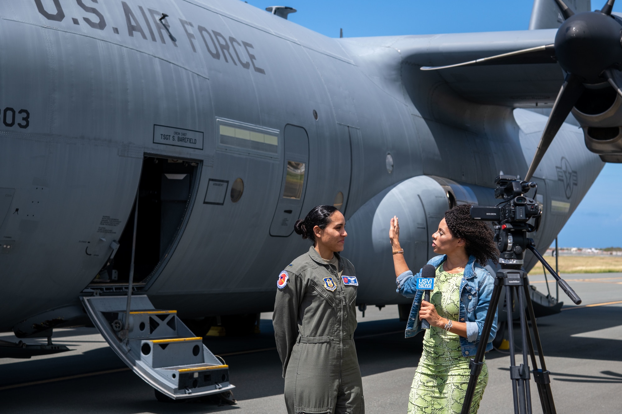WC-130J in the background. A reporter gestures to the aircraft while talking to Lt. Cotto.