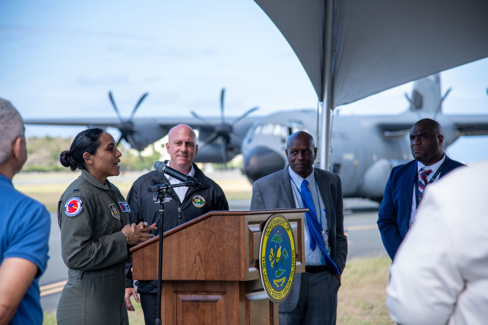 Lt. Cotto stands at a podium speaking. People are in the foreground and background. With a WC-130J in the background also.