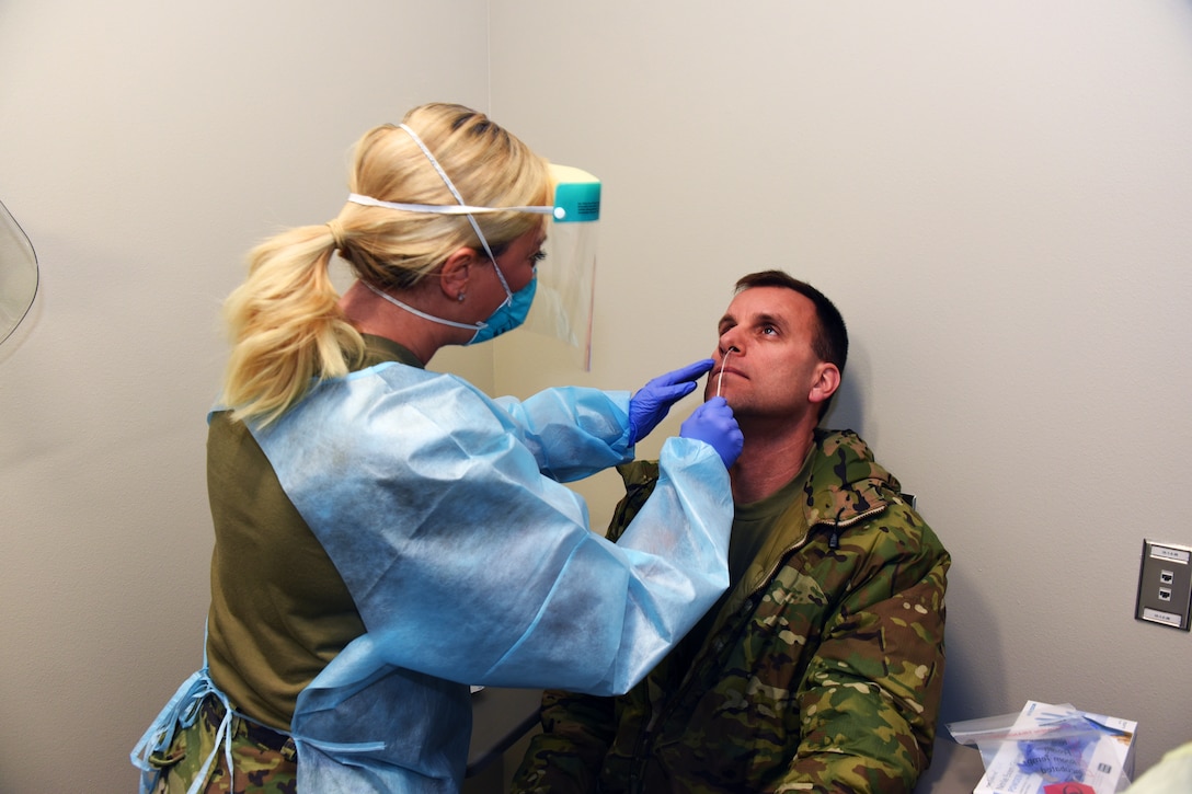 A uniformed woman wearing medical personal protective equipment stands while performing a nasal swab on a seated, uniformed man.