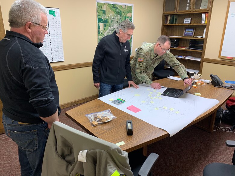 Three men meet in an inside building and examine a map. One of the men is wearing a military uniform.