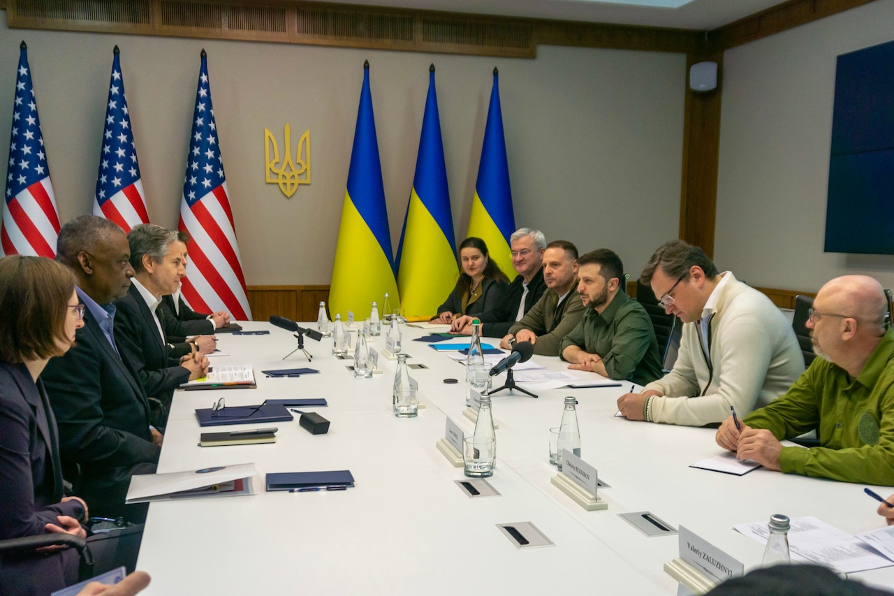 People are seated around a table. United States and Ukrainian flags are in the background.
