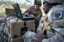 Two soldiers outside work on a laptop computer sitting on a plastic crate.