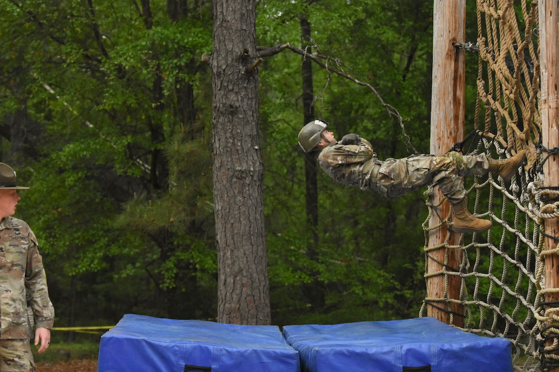 An Army basic trainee climbs up a rope on an obstacle course in the woods as fellow service member watches.
