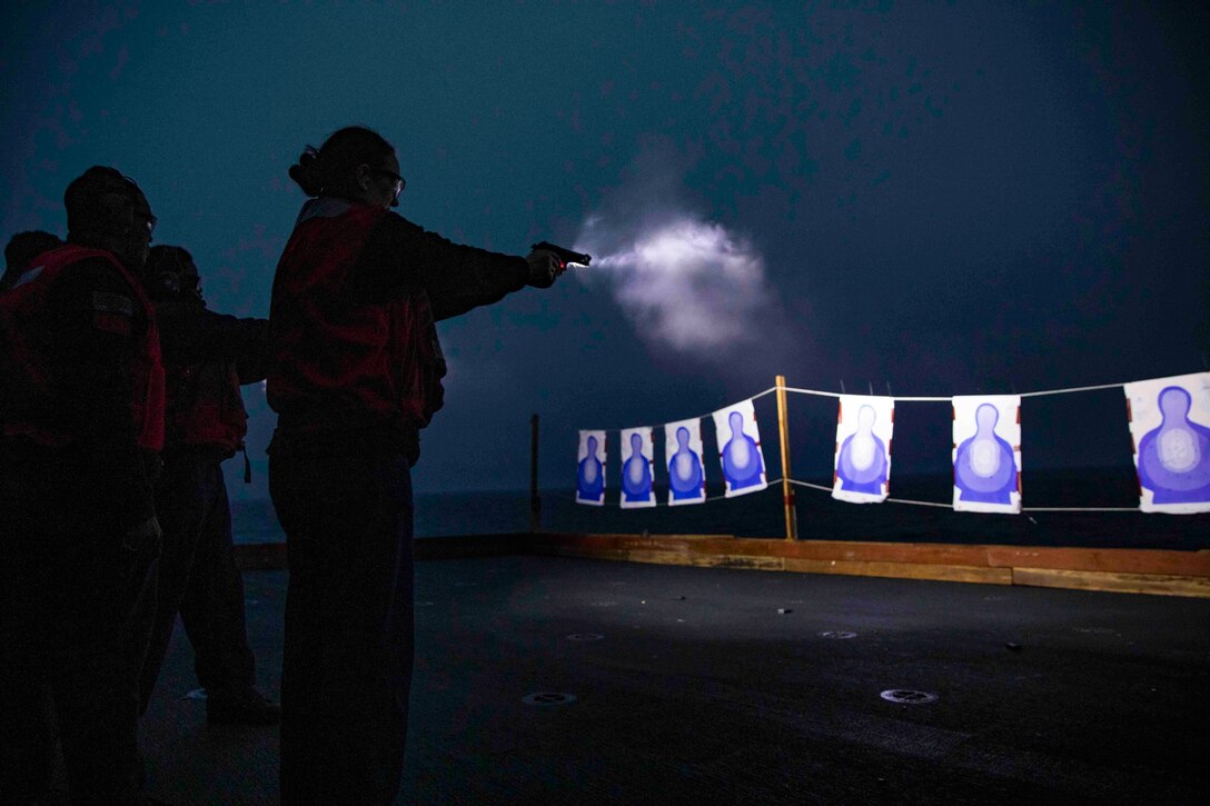 A sailor fires a gun at a target as other stand around.