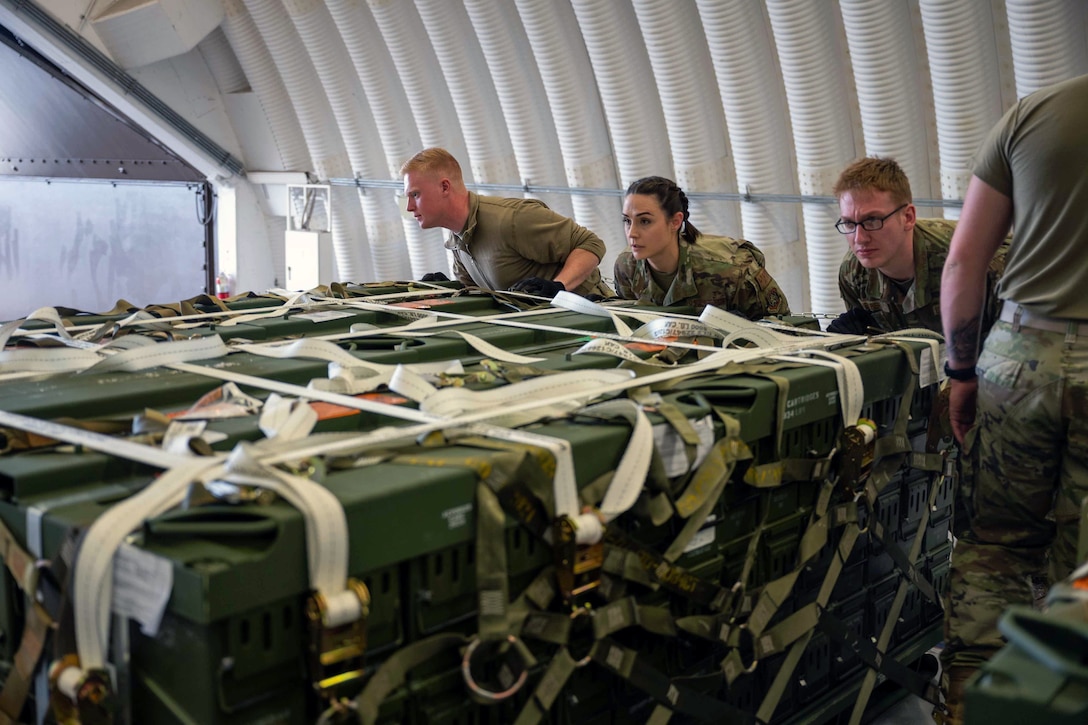 Three Air Force airmen push arrange ammunition cargo pallets.