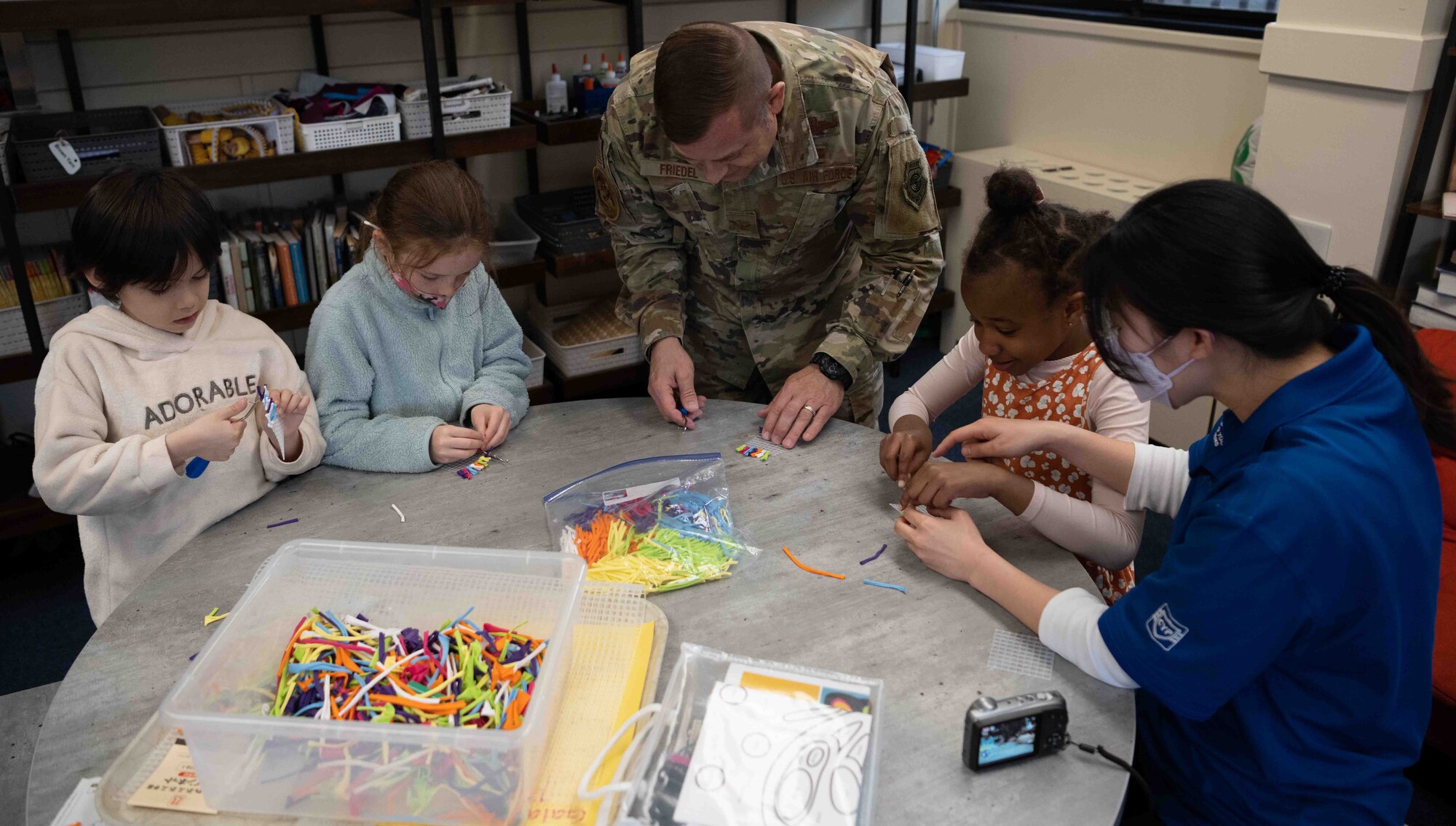 A military member helps craft on a table with three children around him