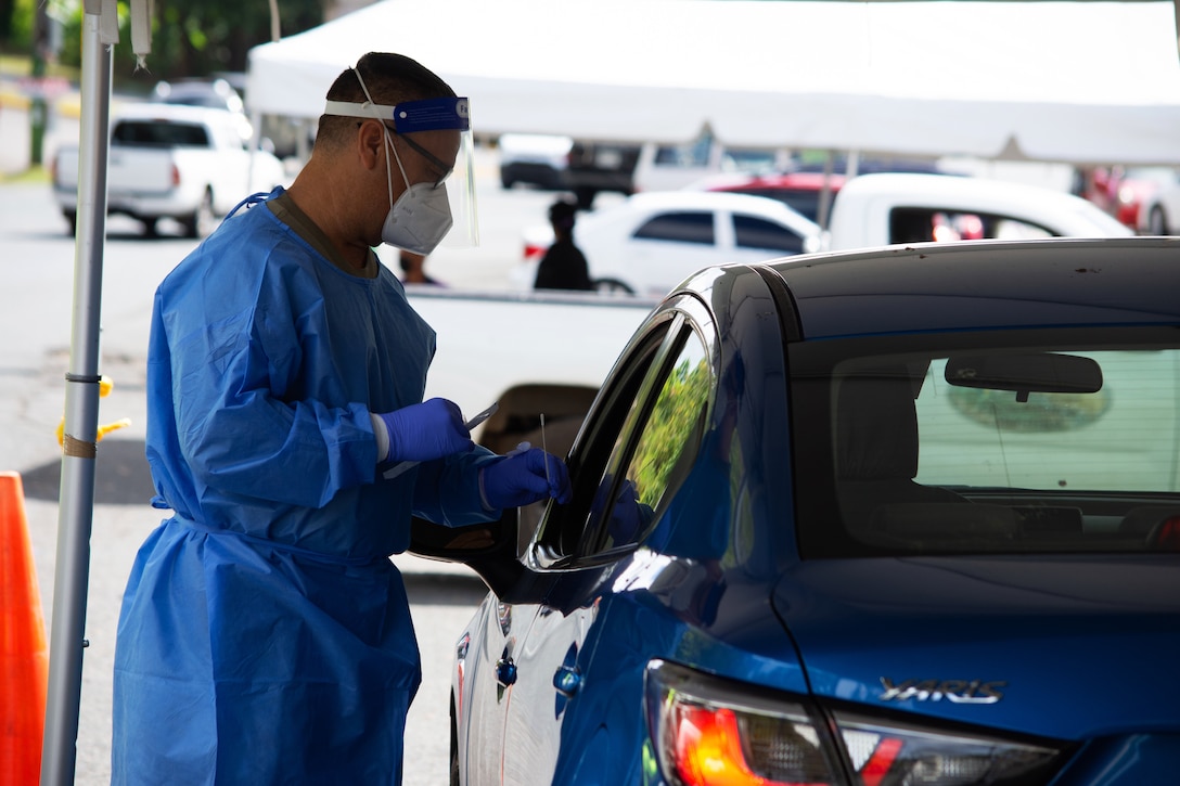A soldier wearing medical personal protective equipment holds a nasal swab while administering a COVID-19 test to someone in their car.