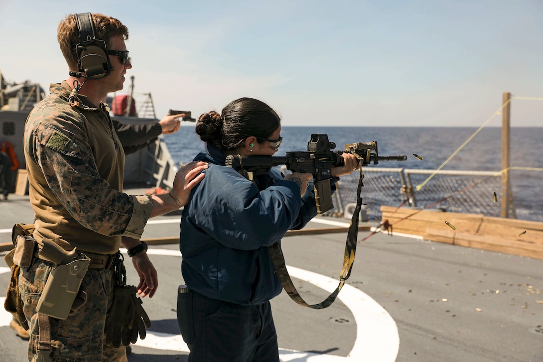 A Marine stands behind a sailor she fires a weapon aboard a ship at sea.