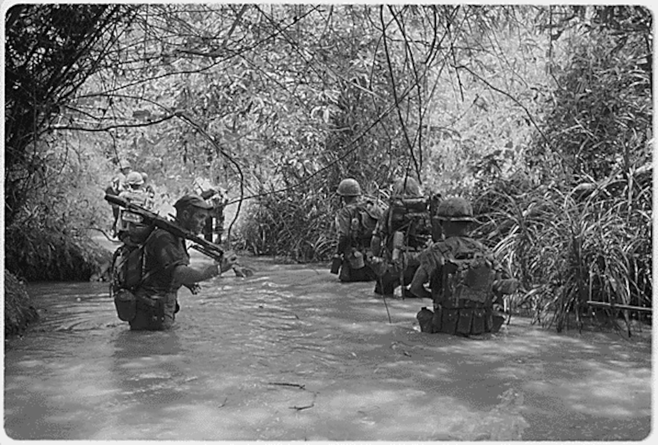 About a half-dozen uniformed men wade through waist-deep water in a jungle.