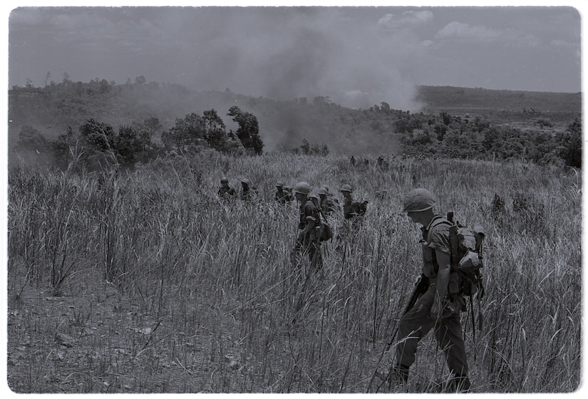 Men in uniform walk through burning fields of tall grass.
