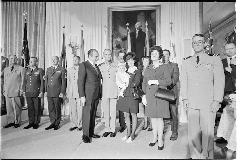 A group of people pose for a photo while service members stand at attention behind them.