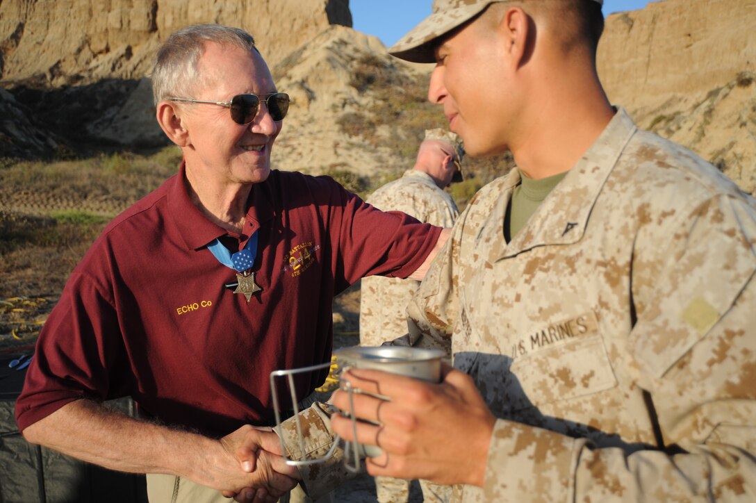 An older man wearing a Medal of Honor shakes the hand of a younger man in uniform amid a mountainous backdrop.