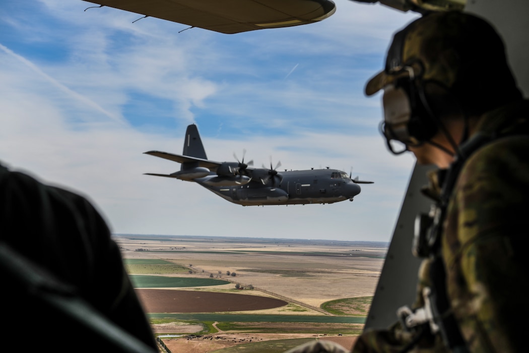 A man wearing a camouflage hat and headphones looks out of a plane towards an MC-103J Commando II aircraft.