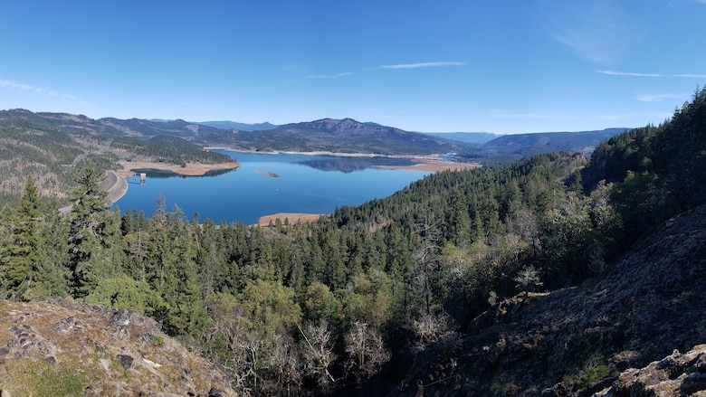 A high-up view of a stunningly blue lake on an equally stunningly sunny afternoon. The lake is surrounded by mountains, hills, and forests of evergreen trees.