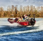 3 people wearing lifejackets on a red boat on a blue water lake with trees in the background.