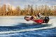 3 people wearing lifejackets on a red boat on a blue lake with trees in the background.