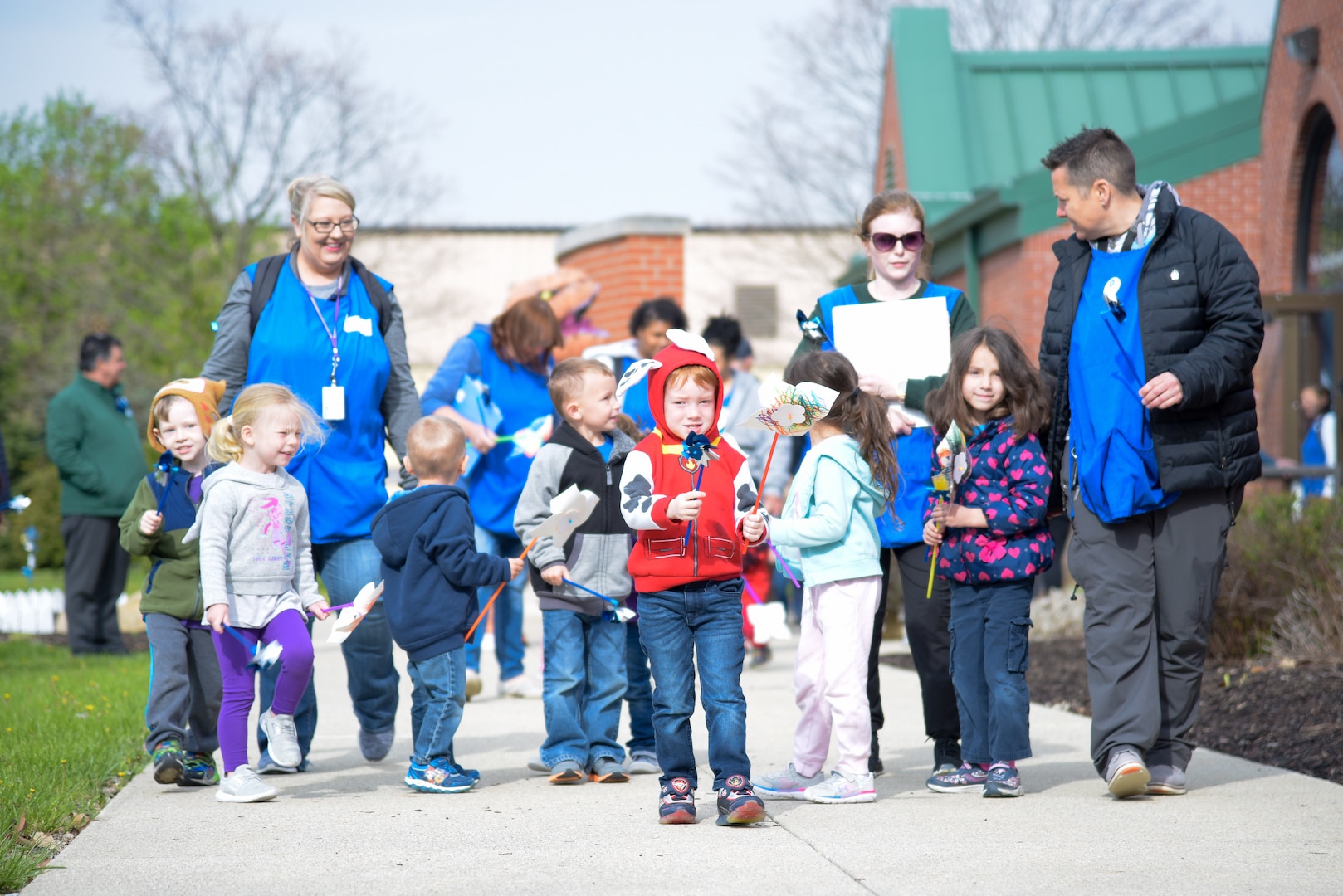 Many children carrying pinwheels march up the sidewalk near the DSCC Child Development Center with the help of blue aproned CDC staff members on a sunny but chilly day.