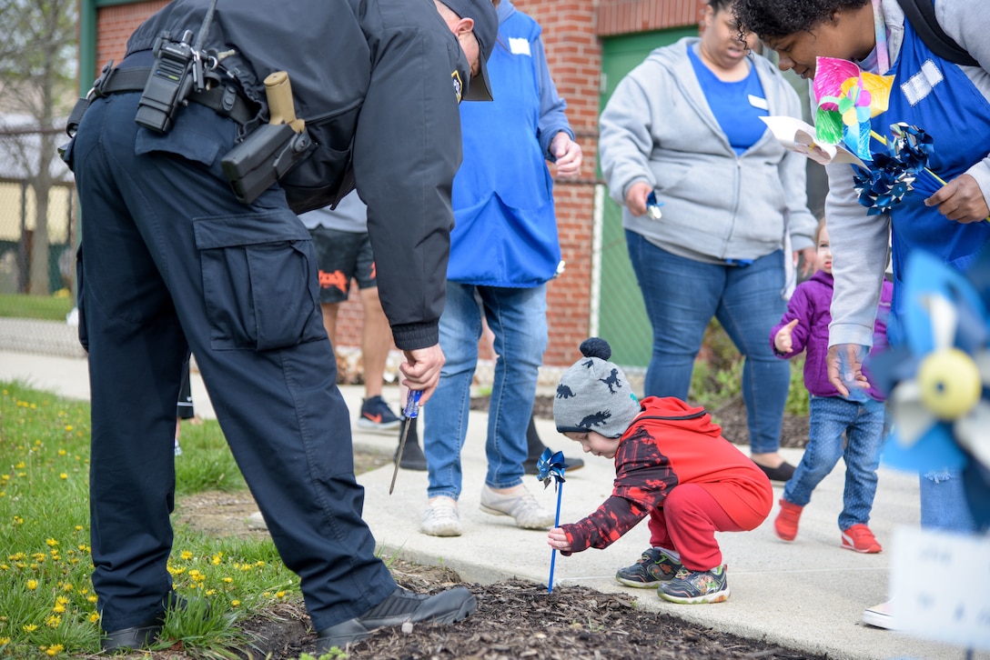 A child in a red suit and grey hat with a pom pom on top squats and places a pinwheel into the ground with the help of a DSCC police officer.