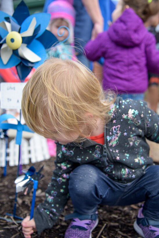 A blond haired child squats and places a pinwheel into the ground.