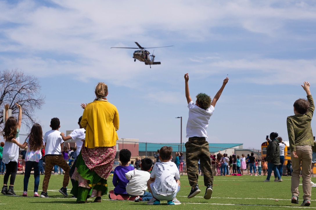 A group of mostly children and adults on a field jump and wave as a helicopter goes by.
