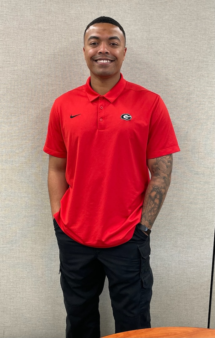 Man in red shirt, black pants standing against grey background