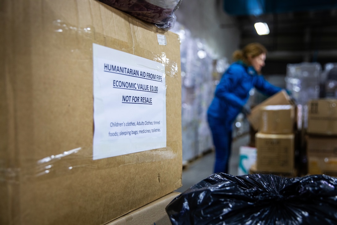 In the foreground, a label with the words “humanitarian aid” is affixed to a box; in the background, a woman lifts a box.