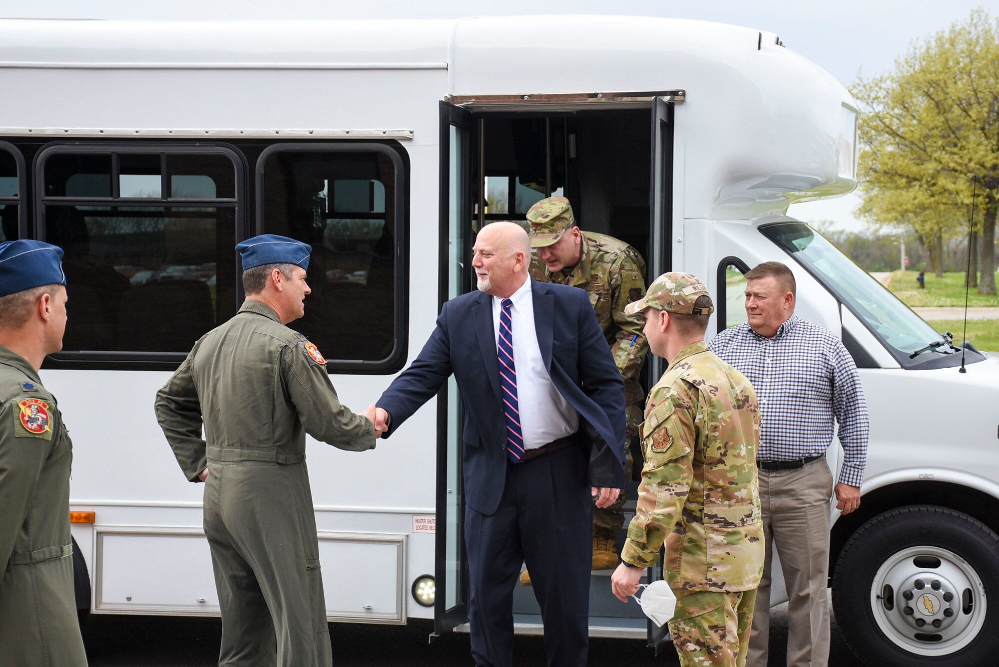 A man in a black suit steps off a bus and shakes hands with a man in a green flight suit.