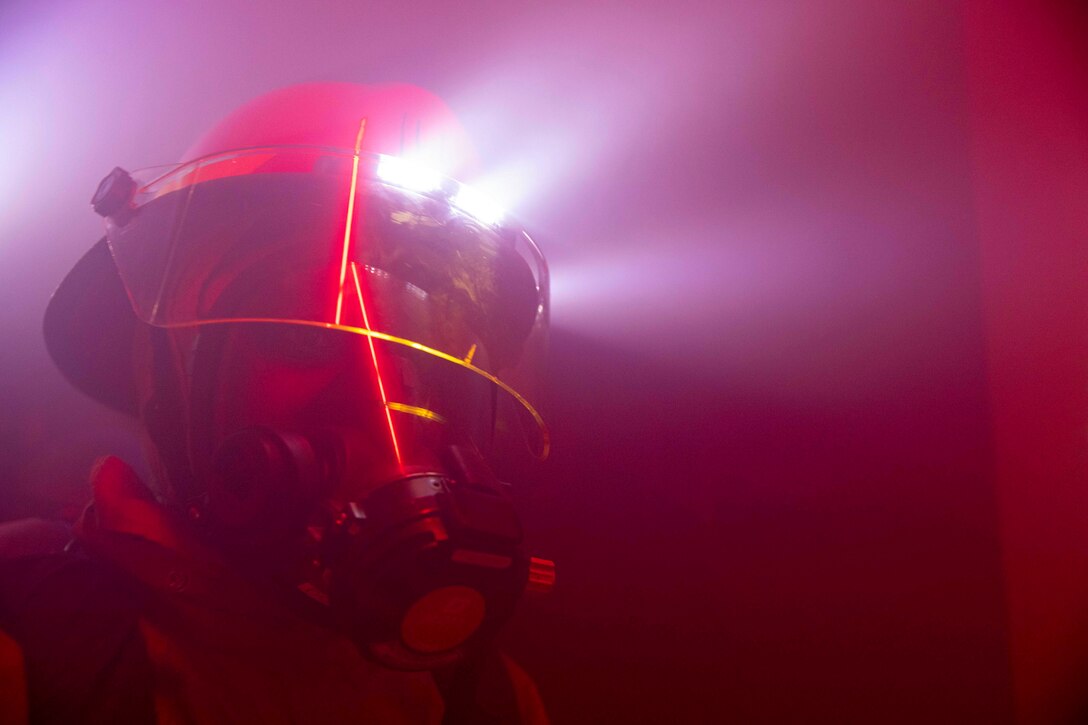 A sailor wearing firefighting gear walks through a dark room.