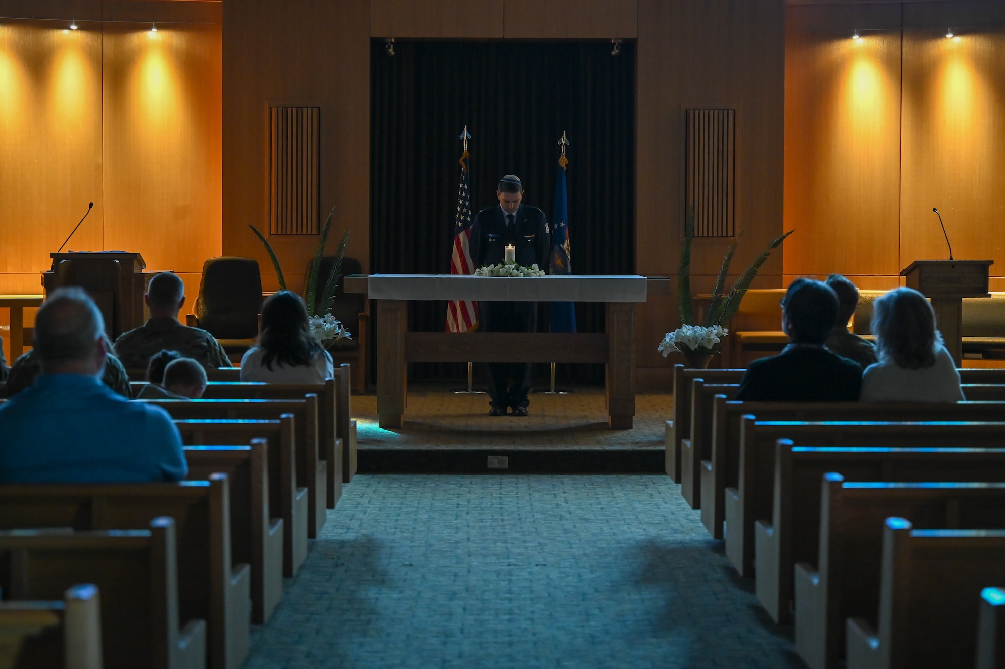 Major Guy Gombo bows his head from behind a lit candle on a table in front of audience members seated in pews.