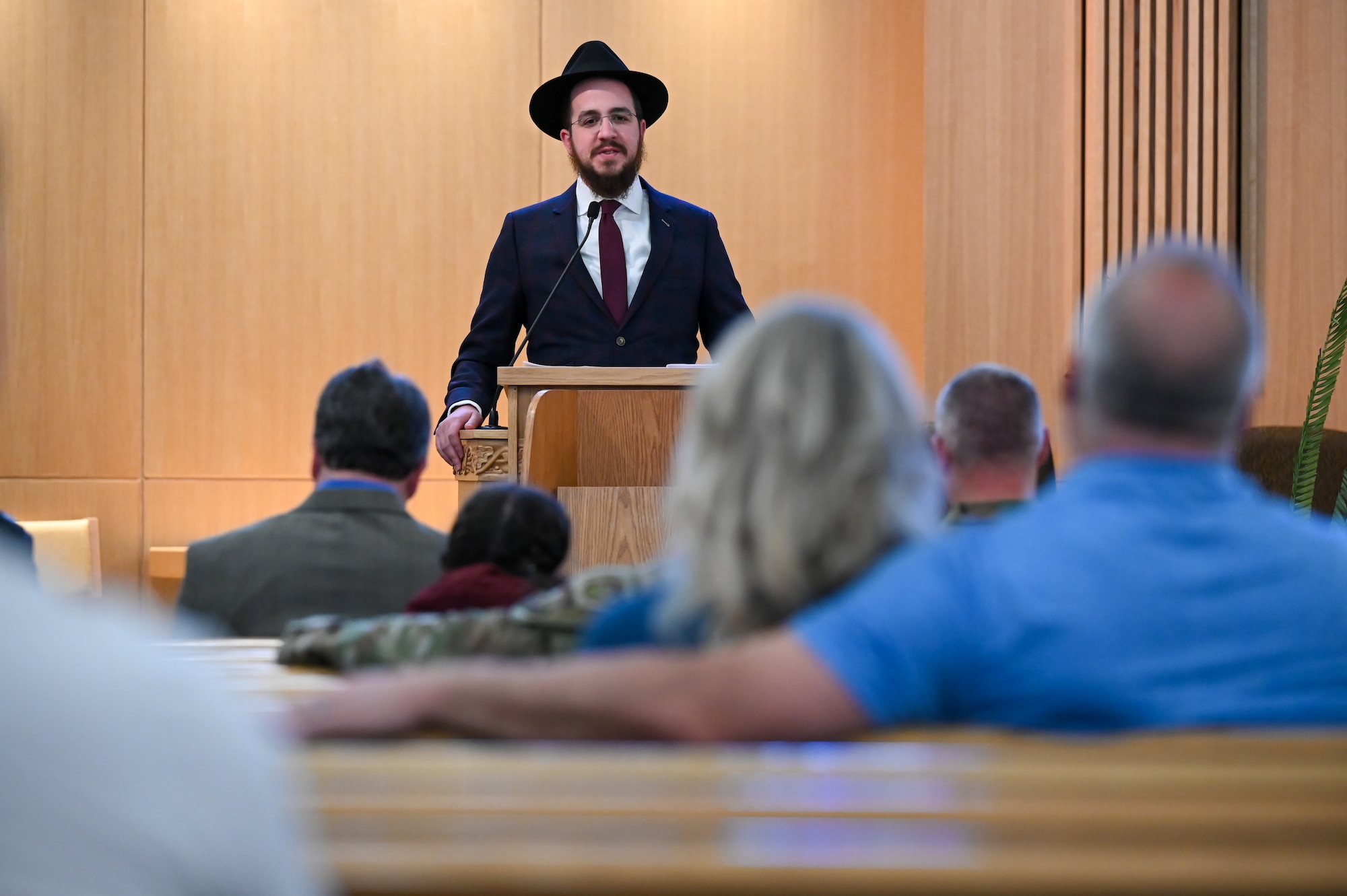 Rabbi Avremi Zippel speaks from behind a podium to audience members seated in pews.