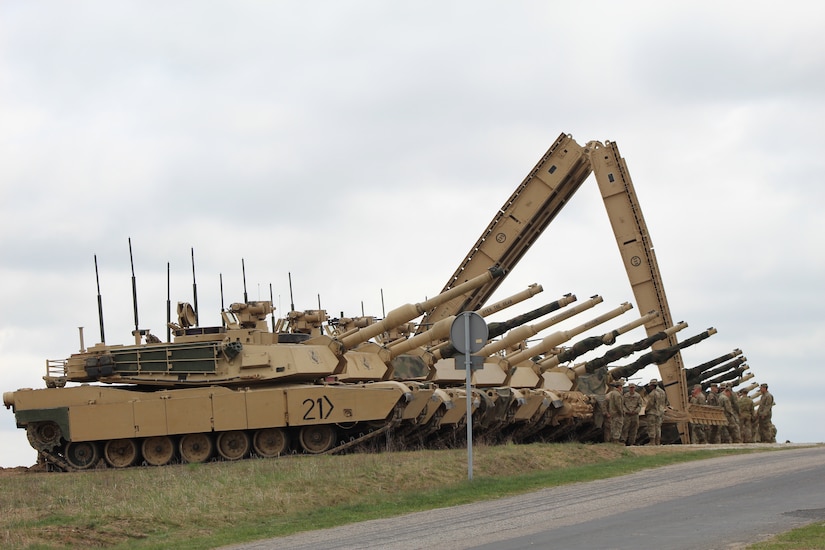 A row of tanks are lined up by the road. A group of service members stand nearby.