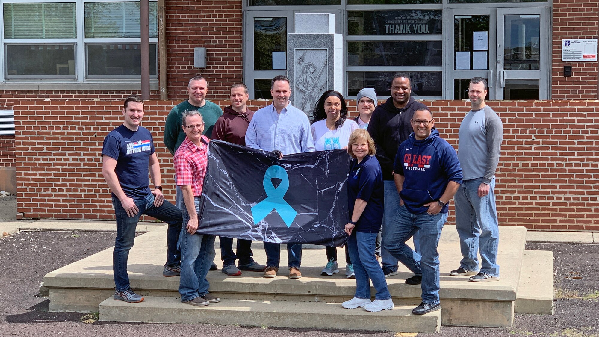 Members of the 111th Attack Wing pose in civilian clothes holding a black banner featuring a teal ribbon in the center.