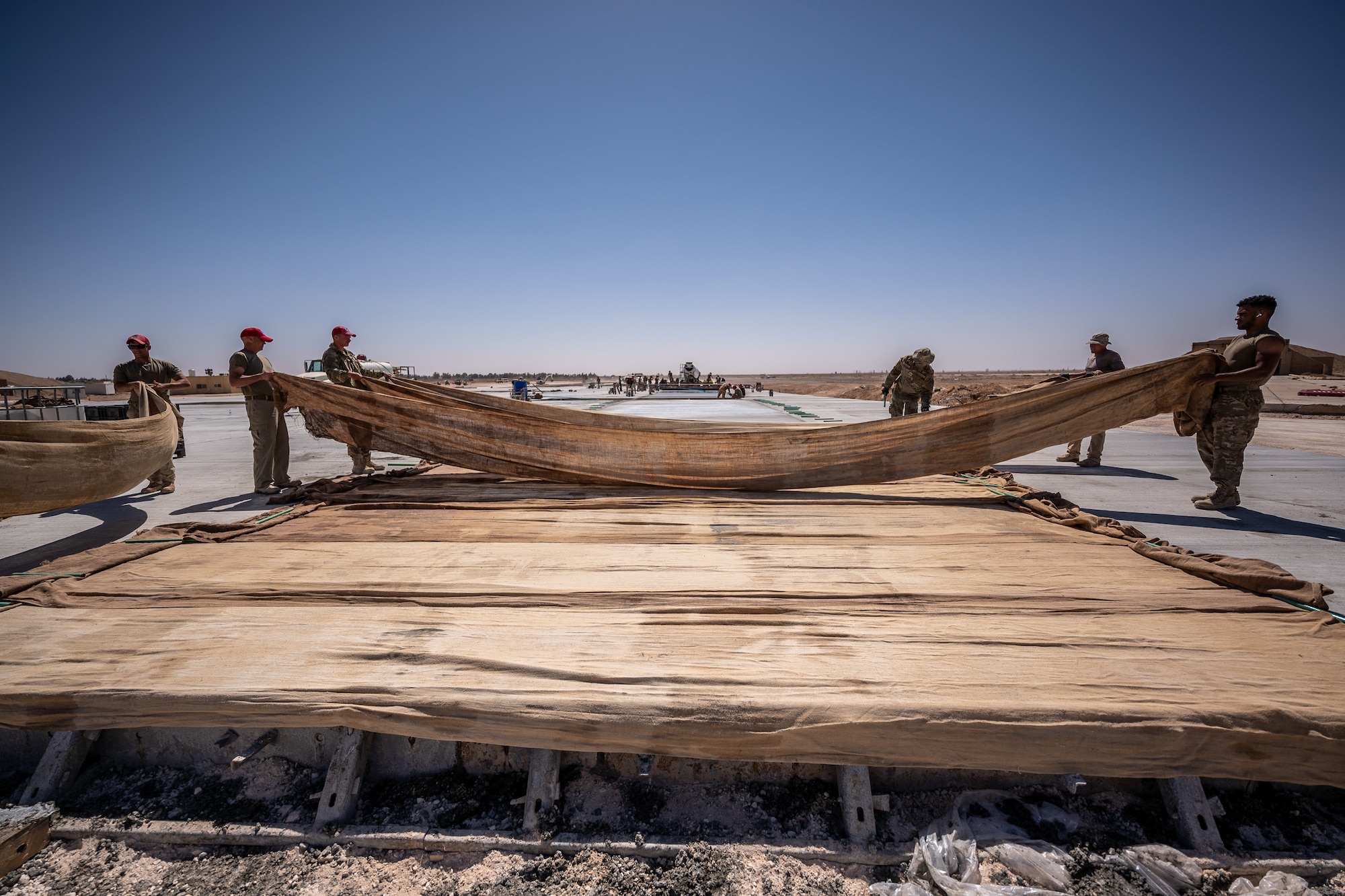 1st Expeditionary Civil Engineer Group Airmen place burlap on top of finished concrete during a runway overrun repair project at an undisclosed location in Southwest Asia, April 14, 2022. Damp burlap cloth is placed on freshly poured concrete and is continuously hosed down to ensure the concrete is hydrated during the curing process.  (U.S. Air Force photo by Master Sgt. Christopher Parr)