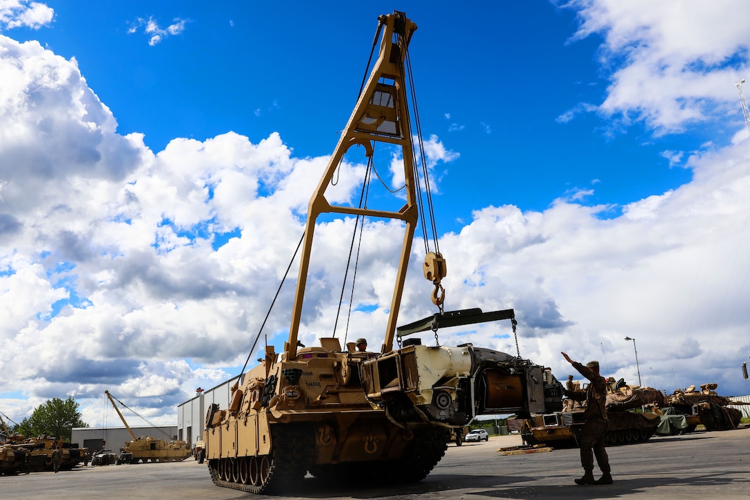 A crane vehicle with tank tread lifts a piece of a equipment while a soldier stands nearby and provides direction to the driver.