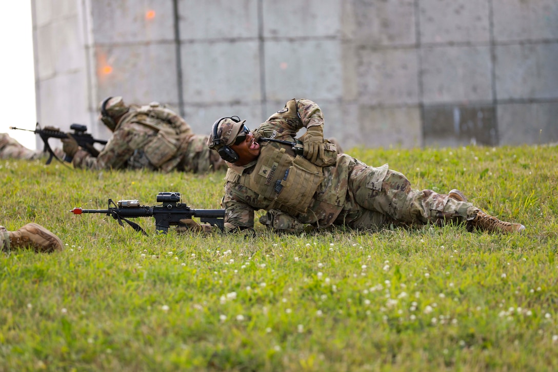 A soldier lying on the ground with a weapon yells at someone.