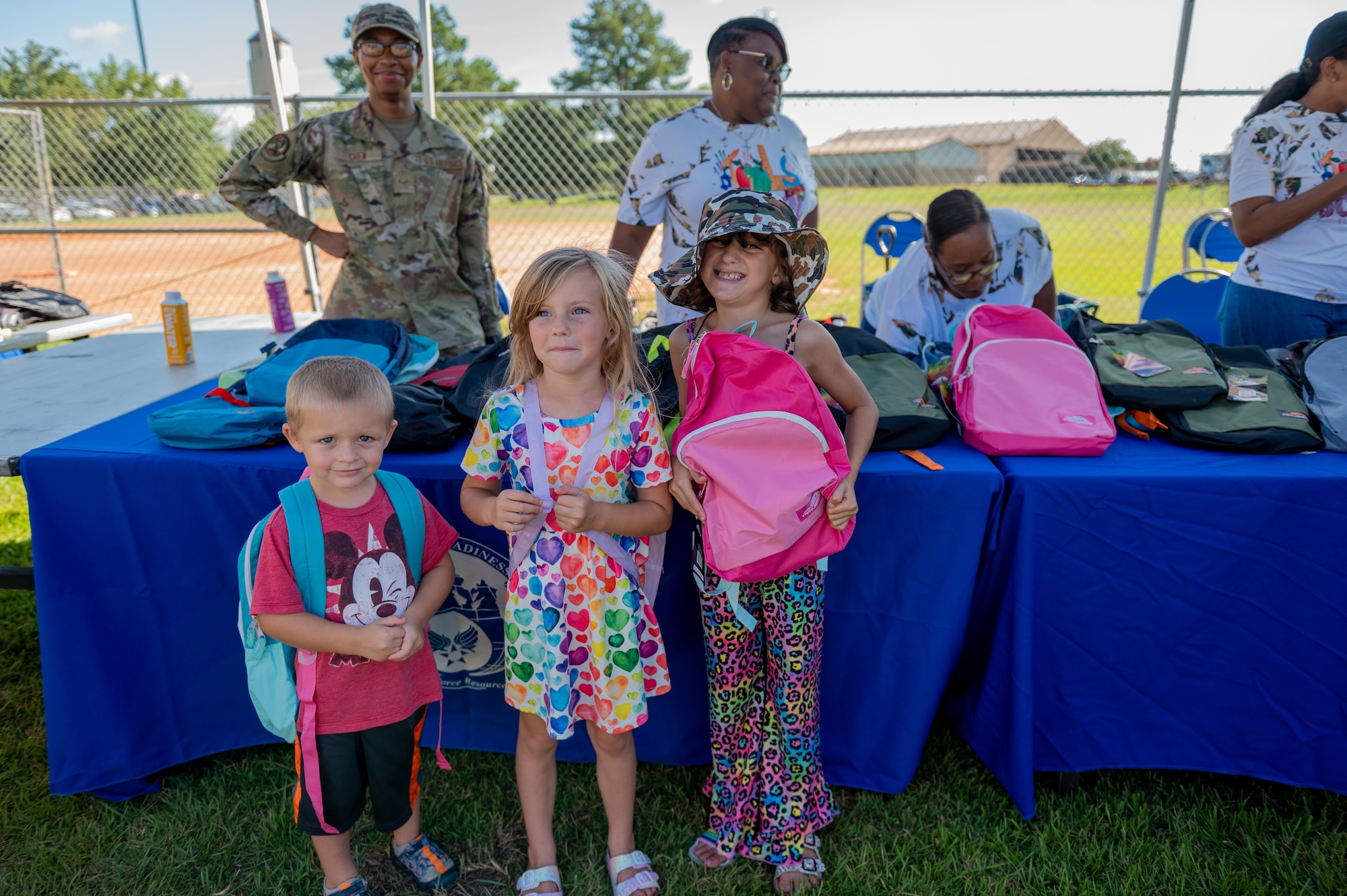 Children stand together under an outdoor tent while holding backpacks.