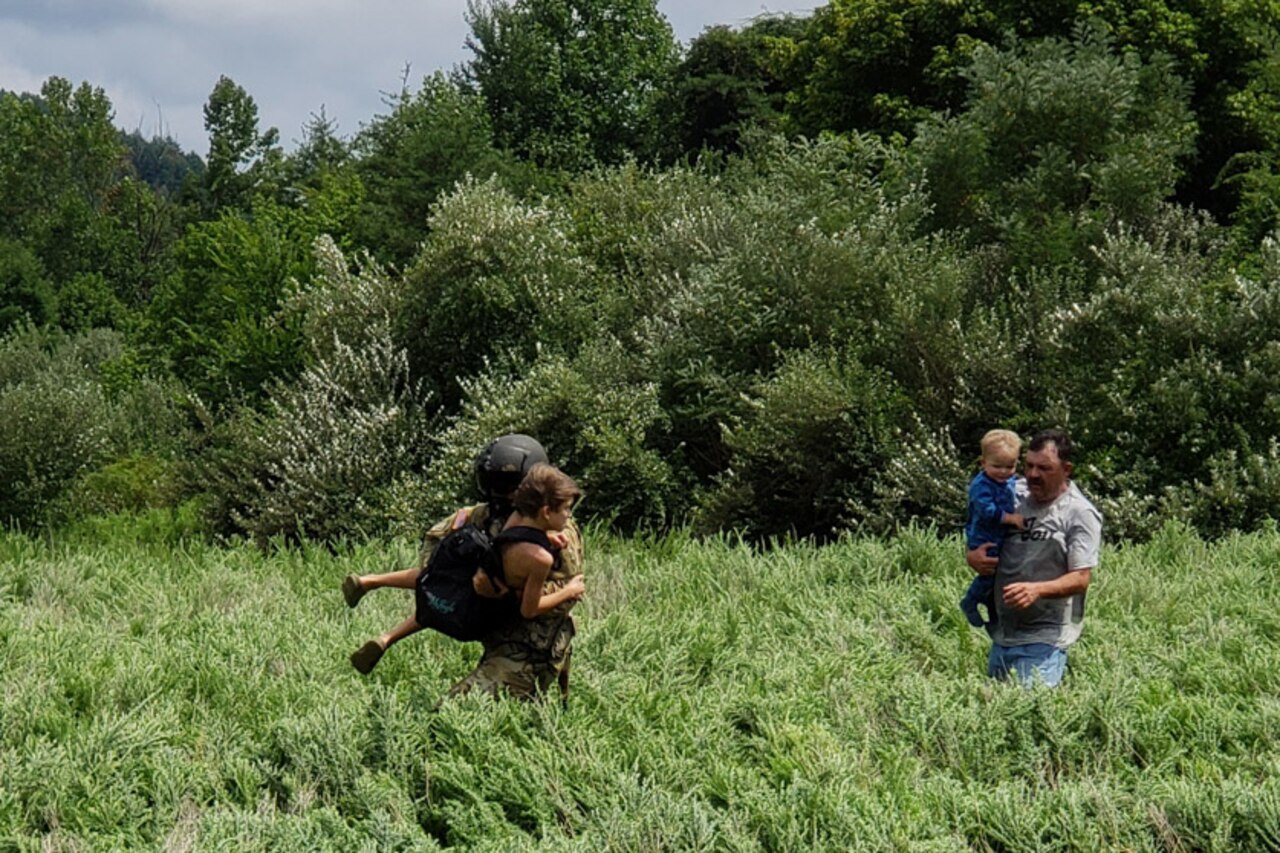 A Kentucky National Guardsman carries a civilian through tall grass, as another civilian carrying a toddler follows on foot.