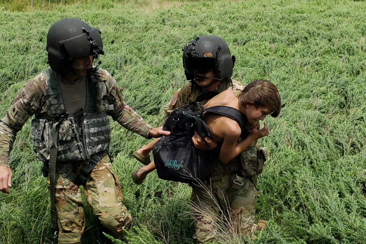 Two service members carry a child through a field.