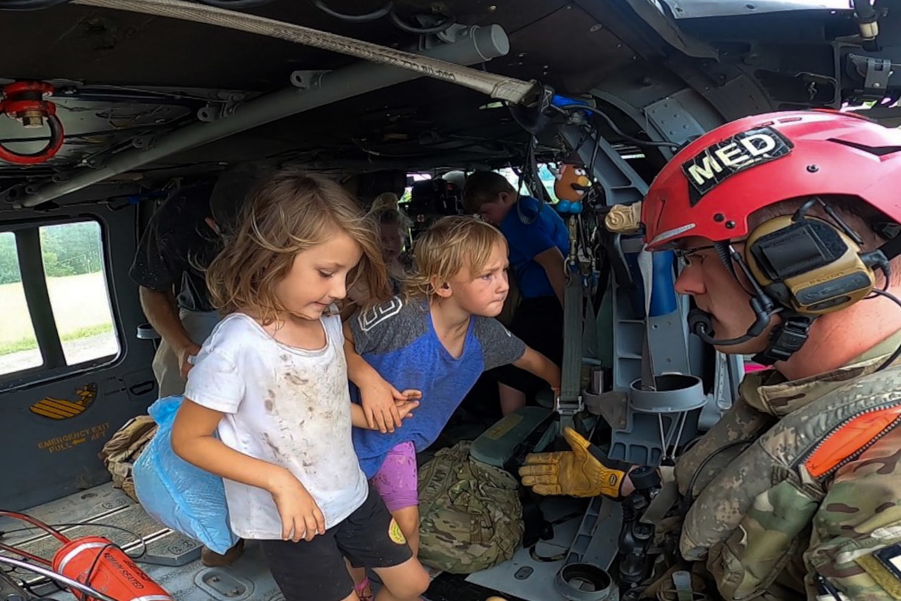 A service member holds his hand out toward two little children on an aircraft holding hands.