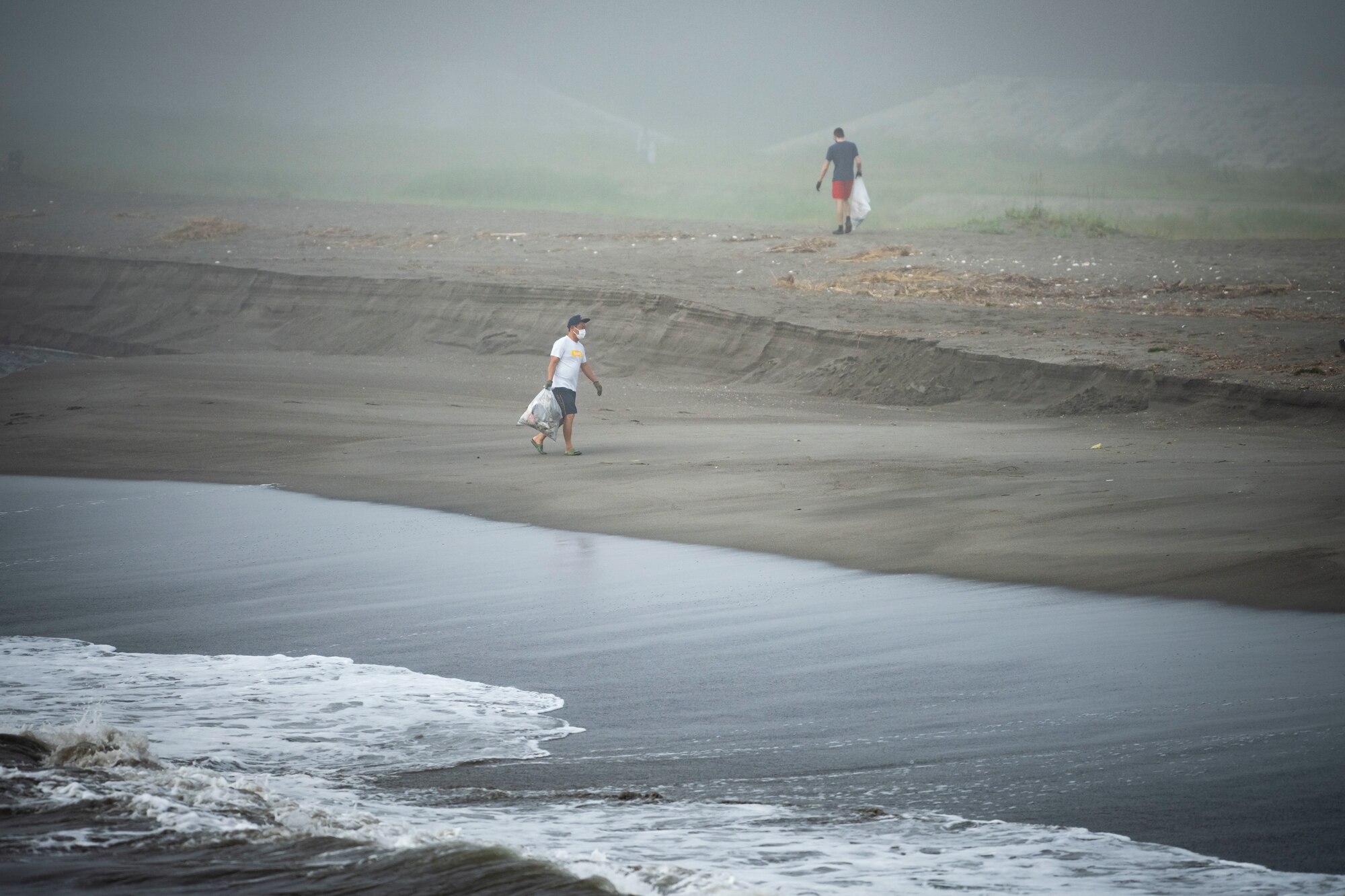 A Team Misawa member holds garbage bags while walking next to waves crashing.