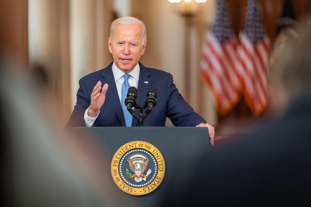 President Joe Biden gestures with his hand while speaking from a lectern with flags in the background.