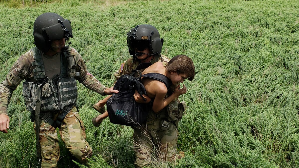 Two National Guardsmen walk through tall grass, one carrying a small person.